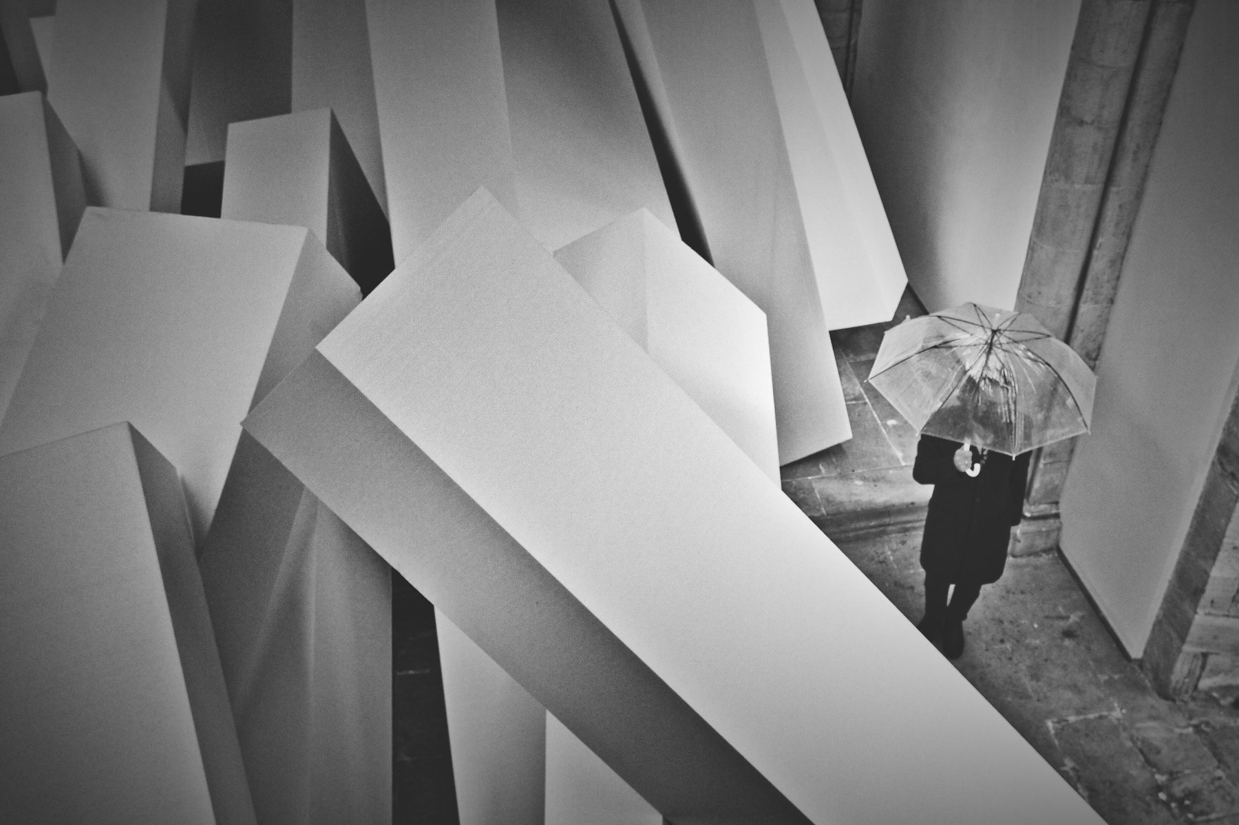 Black and white photo of a person with an umbrella walking past angular paper sculptures